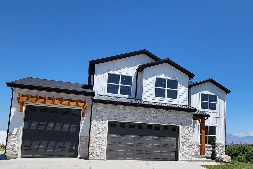 Modern two-story house with a stone and white siding exterior, black roof, large windows, and three dark garage doors. A wooden pergola is above one garage. Residential Construction Services Utah crafted this home under a clear blue sky.