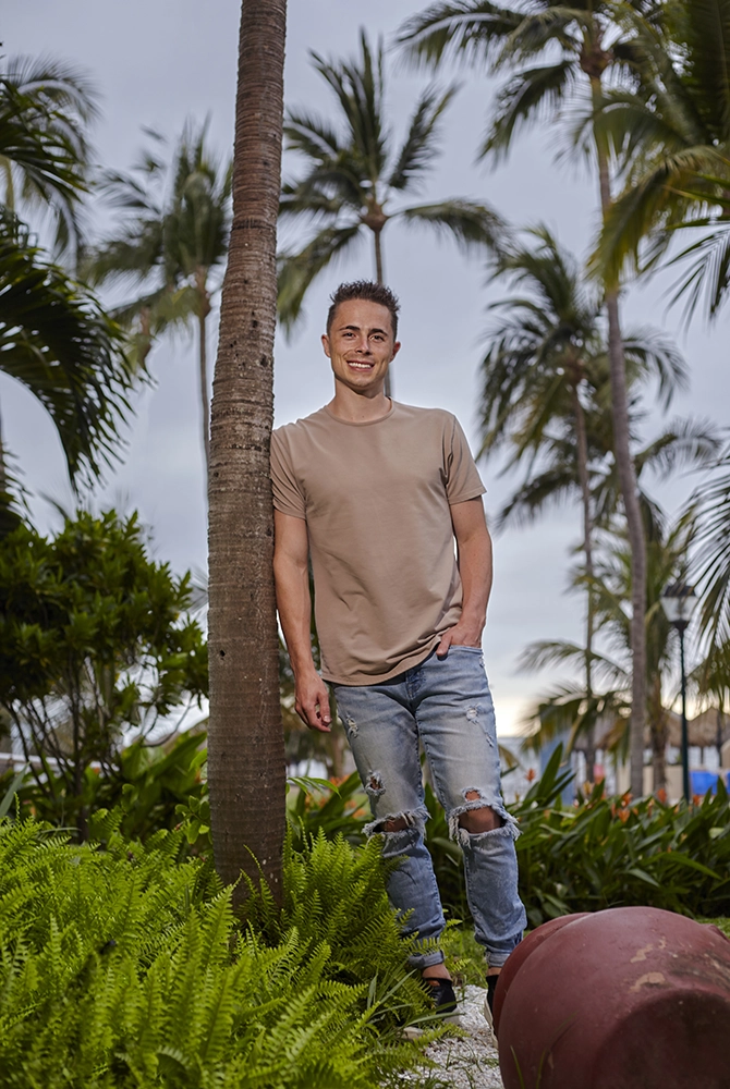 A man wearing a beige t-shirt and ripped jeans leans against a palm tree, smiling, surrounded by lush green plants. He looks relaxed beneath the cloudy sky, taking a break from his job at a construction company in Utah.