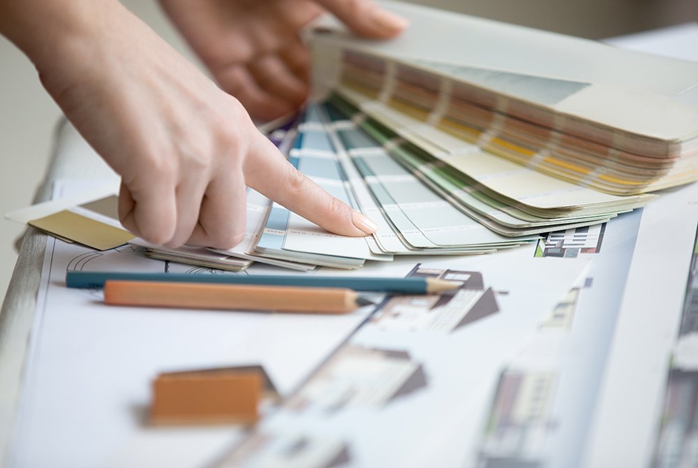 A person’s hand flipping through a palette of paint color swatches over architectural drawings, with pencils and a ruler nearby—capturing the thoughtful planning behind quality Residential Construction Services Utah.
