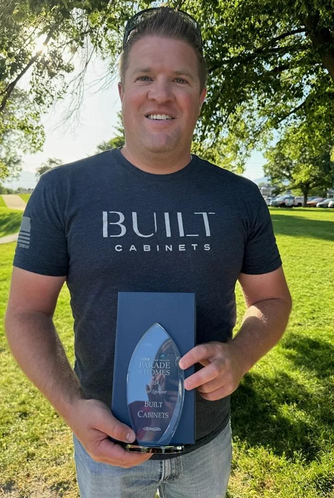 A man in a "BUILT CABINETS" t-shirt stands outdoors on grass, smiling and holding a glass award that says "Parade of Homes 2023" and "BUILT CABINETS," representing a top construction company in Utah. Trees and sunlight brighten the background.