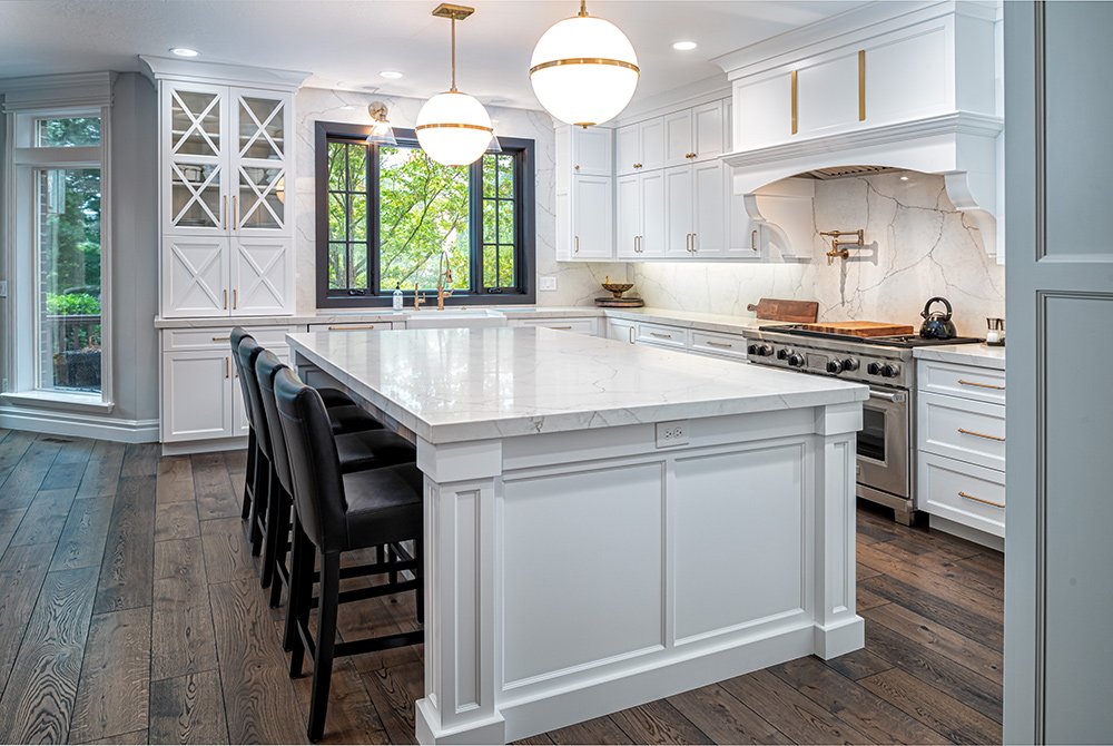 Modern kitchen with custom cabinetry Utah, white cabinets, a large marble island with black chairs, dark wood floors, pendant lights, and a window overlooking greenery. Stainless steel appliances and glass cabinet doors are visible.