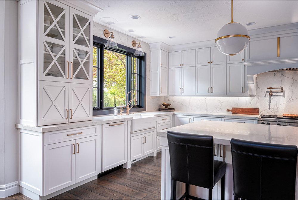 Bright, modern kitchen with white cabinets, marble countertops, a large island with two black chairs, gold accents, and dark wood floors—expertly crafted by our Residential Construction Services Utah team.