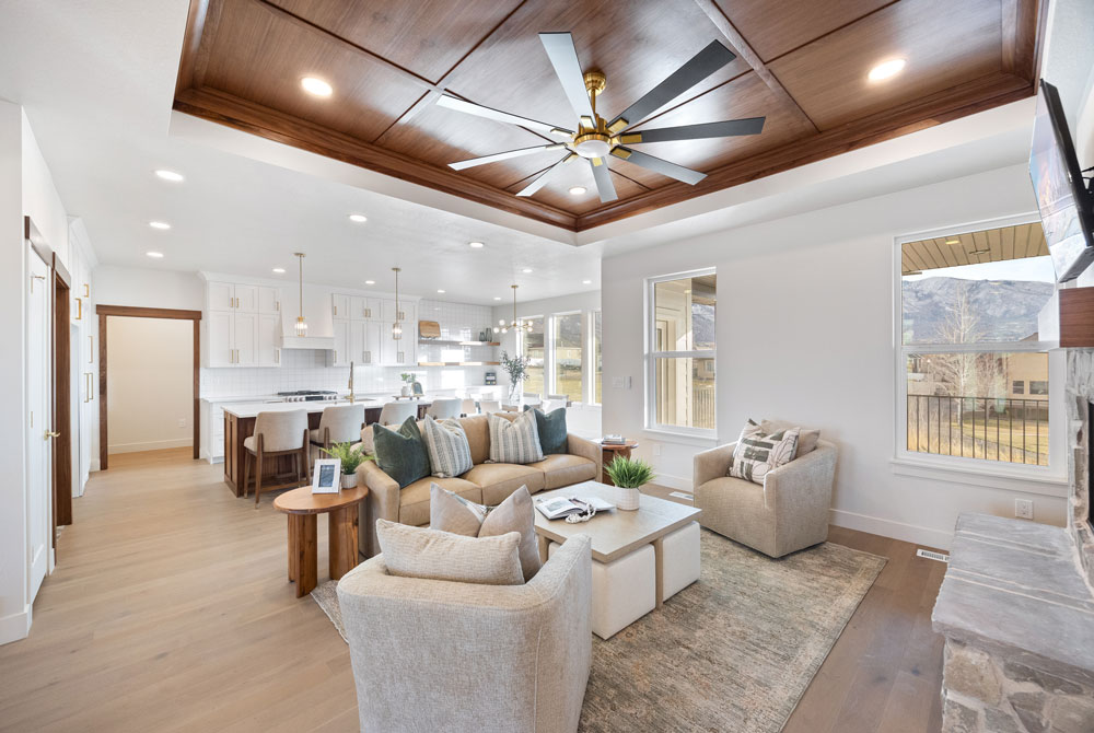 Bright, modern open-concept living room and kitchen with light wood floors, beige furniture, a coffered ceiling with a large fan, and large windows showing mountain views. Contact Home Builders in Utah to bring this stunning vision to life.