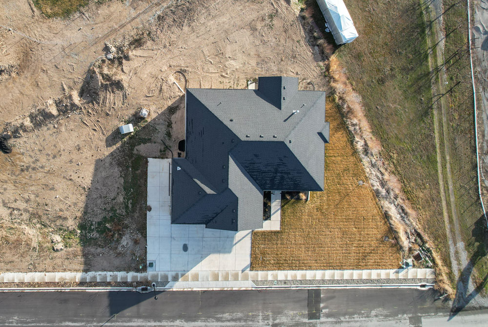 Aerial view of a modern house with a gray roof, concrete driveway, and small patch of lawn bordered by a sidewalk—an example of quality Residential Construction Services Utah—surrounded by dirt and sparse grass on a clear day.
